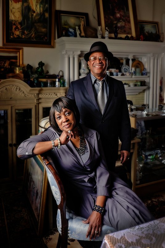 Photo of a black woman entrepreneur sitting on a blue antique chair, with her black professional husband standing behind her. 