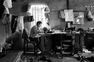 A Myanmar tailor sews a traditional outfit in her shop. Photograph in black and white. 