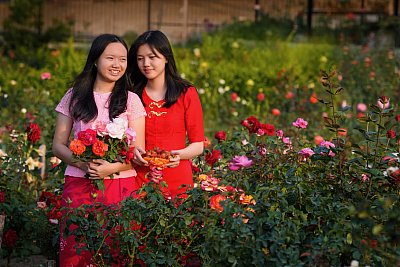 Photograph of two women in traditional Myanmar clothes holding flowers and strawberries in a rose garden. 
